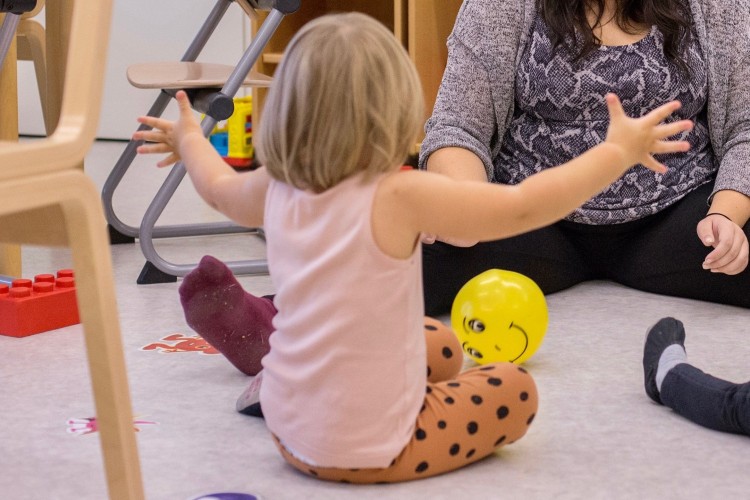 Ett barn sitter på golvet och rullar en boll mot en vuxen. Barnet har ryggen mot kameran.