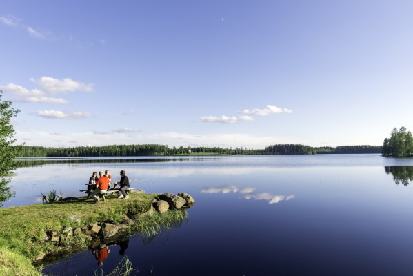 Rauhallinen kesäkuva järven rannalta. Muutamia henkilöitä istuu penkillä rannalla.