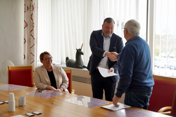 Kommunstyrelsens ordförande Johanna Holmäng sitter vid ett bord och ser på när kommundirektören Stefan Svenfors skakar hand och tar emot papper av Henrik Sandberg, ordförande för Esse pensionärsförening.